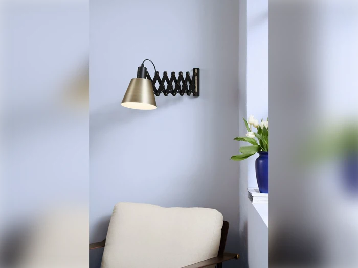 En vægmonteret lampe i messing med harmonika-arm lyser over en beige lænestol; en blå vase med hvide tulipaner står i en nærliggende vindueskarm.