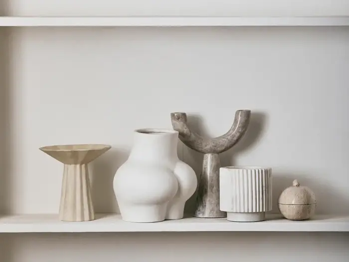 Fem moderne keramikvaser og -kar i forskellige abstrakte former og neutrale toner står på en hvid hylde mod en lys væg.