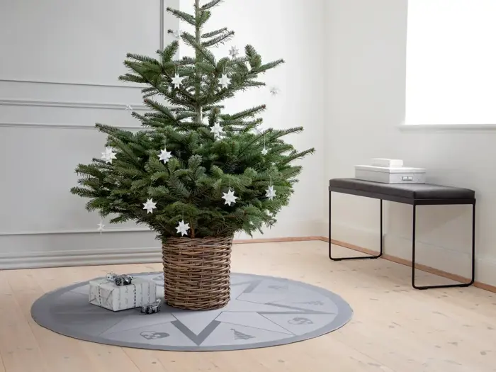 Et juletræ i en flettet kurv, pyntet med hvide stjerneornamenter, står på et gråt træskørt med gaver i nærheden i et minimalistisk rum.