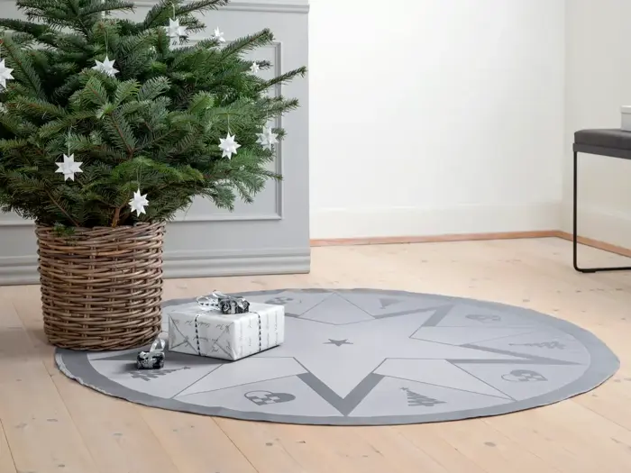 Et pyntet juletræ i en flettet kurv står på et trægulv ved siden af en indpakket gave og et rundt, stjernemønstret gråt træskørt.