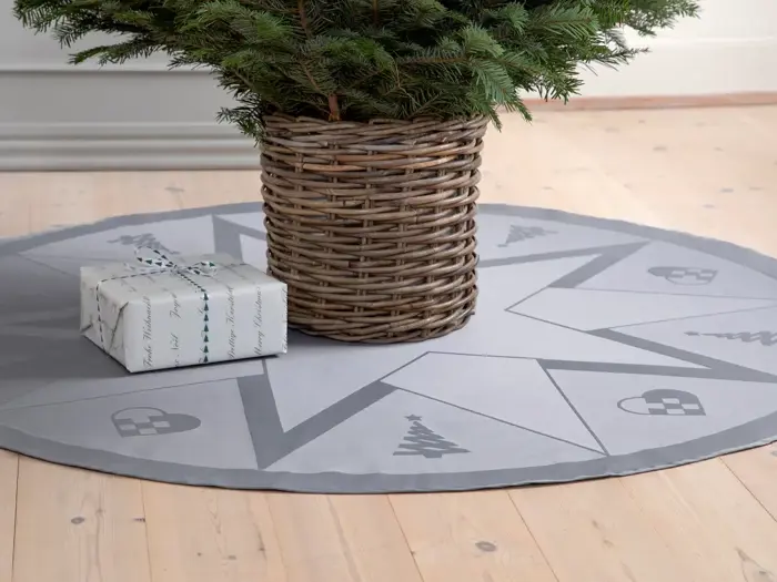 En flettet kurv med et juletræ står på et rundt gråt træskørt med stjerne- og julemotiver; en indpakket gave ligger ved siden af på et lyst trægulv.