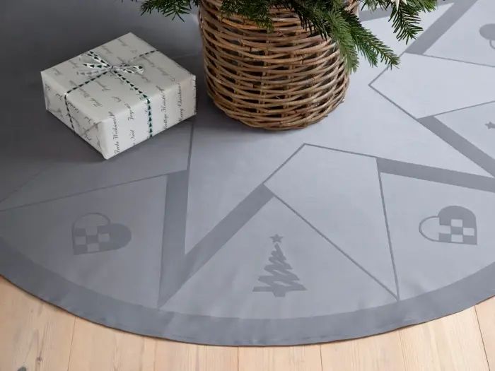 En indpakket gave ligger på et lyst trægulv ved siden af et rundt gråt træskørt med diskrete julemotiver, bl.a. et træ og hjerter. En flettet kurv med fyrretræsgrene står på skørtet.
