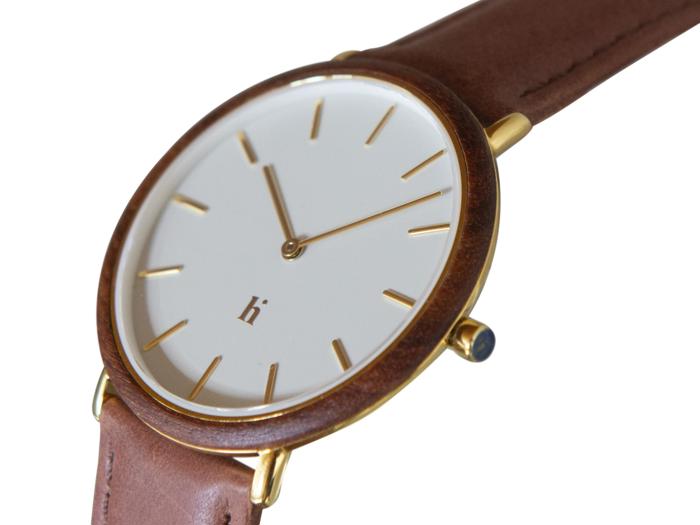 Et minimalistisk armbåndsur med hvid urskive, timemarkeringer og visere i guld, en brun læderrem og en urkasse i guld. Tiden viser ca. 10:10.