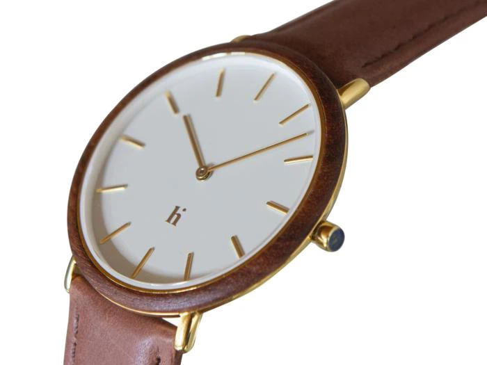 Et minimalistisk armbåndsur med hvid urskive, timemarkeringer og visere i guld, en brun læderrem og en urkasse i guld. Tiden viser ca. 10:10.
