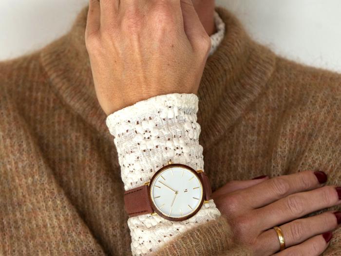 En person iført en solbrun sweater og et hvidt hæklet ærme viser et guldkantet armbåndsur med en brun læderrem på sit håndled.