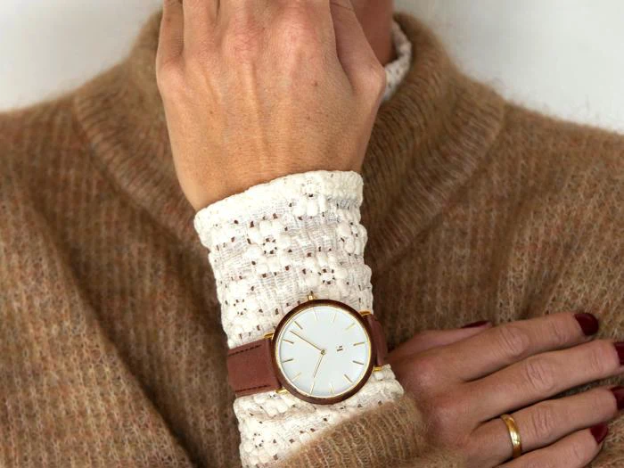 En person iført en solbrun sweater og et hvidt hæklet ærme viser et guldkantet armbåndsur med en brun læderrem på sit håndled.