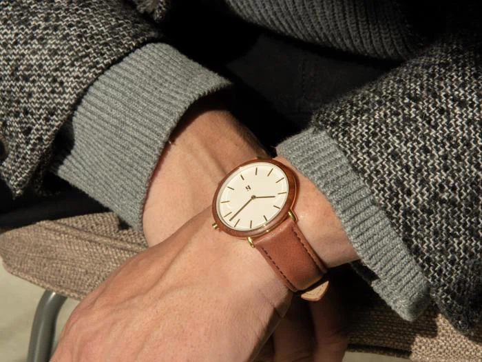 En person iført grå sweater og mønstret frakke har et brunt læderarmbåndsur med hvid urskive og gulddetaljer på venstre håndled.