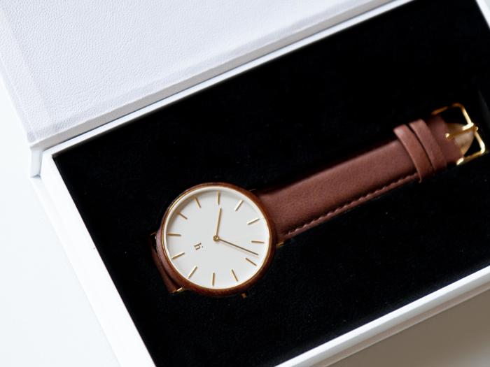 Et minimalistisk armbåndsur med brun læderrem og hvid urskive i en sort og hvid præsentationsæske.