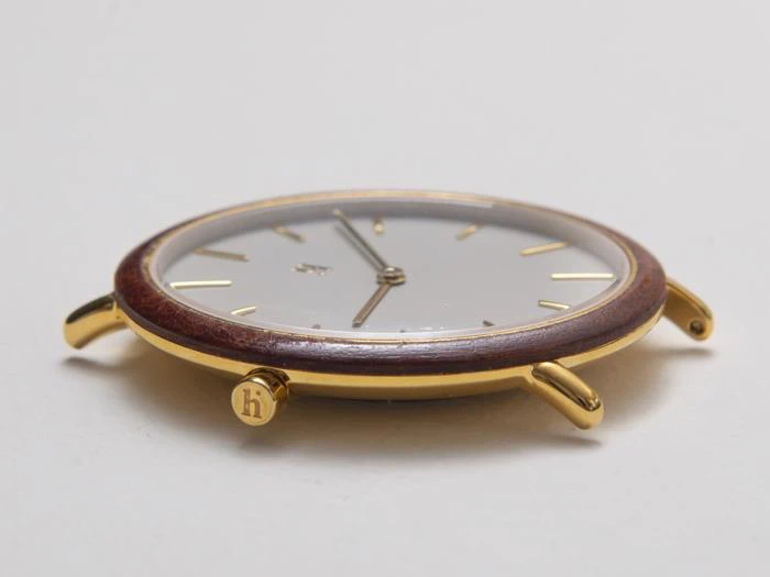 Et nærbillede fra siden af et tyndt, guldfarvet og brunt armbåndsur med en minimalistisk hvid urskive og enkle timemarkeringer, der ligger fladt på en hvid overflade.