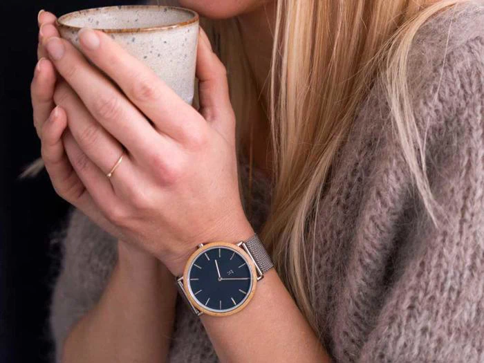 En kvinde i en lodden grå sweater holder et keramikkrus med begge hænder og bærer et sølvfarvet armbåndsur med en mørk urskive og en tynd guldring på fingeren.