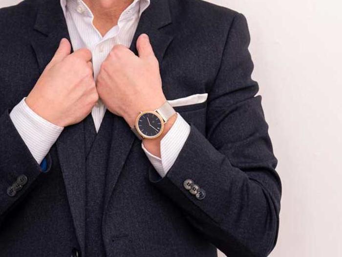 En mand i mørkt jakkesæt retter på sin jakke, han er iført en hvid skjorte og et armbåndsur med sort urskive og guldkant.