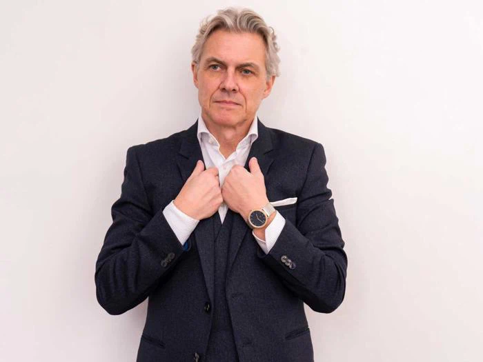 En midaldrende mand med gråt hår i mørkt jakkesæt retter på sin jakke, mens han står på en almindelig hvid baggrund og kigger lidt til siden.