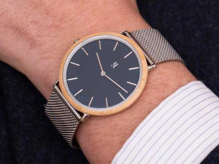 En person bærer et minimalistisk armbåndsur med sort urskive, sølvfarvet mesh-rem og lys træramme på sit venstre håndled.