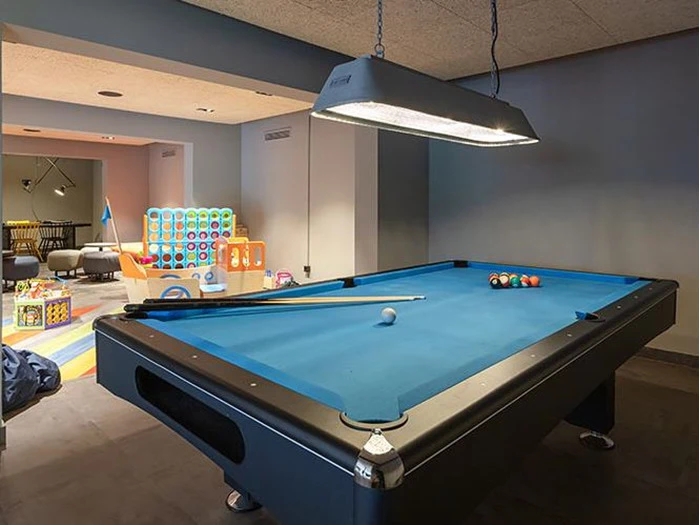 Et blåt poolbord med køer og bolde står i et rum med børnelegetøj, bl.a. en kæmpe Connect Four, på farverige måtter i baggrunden.