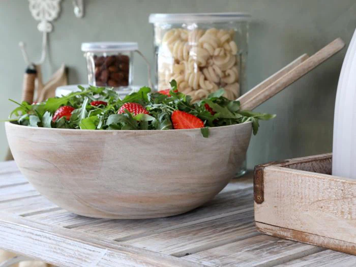 En træskål med salat med rucola og jordbær i skiver står på et rustikt bord med krukker med pasta og nødder i baggrunden.