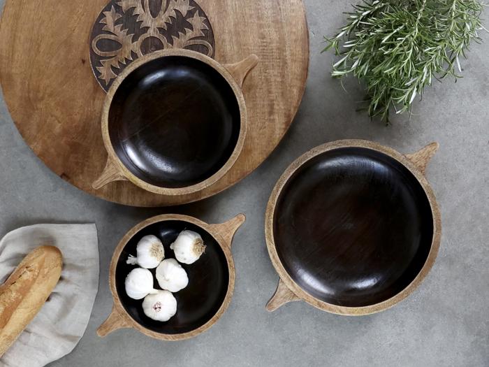 Tre træskåle, den ene fyldt med hvidløgsfed, er anbragt på en grå flade med et træbræt, en rosmarinkvist og en baguette på en serviet ved siden af.