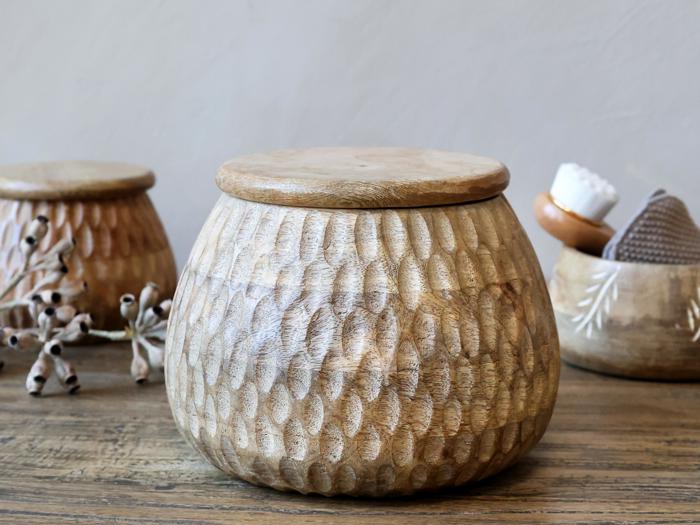 En rund, struktureret træbeholder med låg står på en træoverflade. I baggrunden er der lignende beholdere, en gren med bælge og plejeartikler.