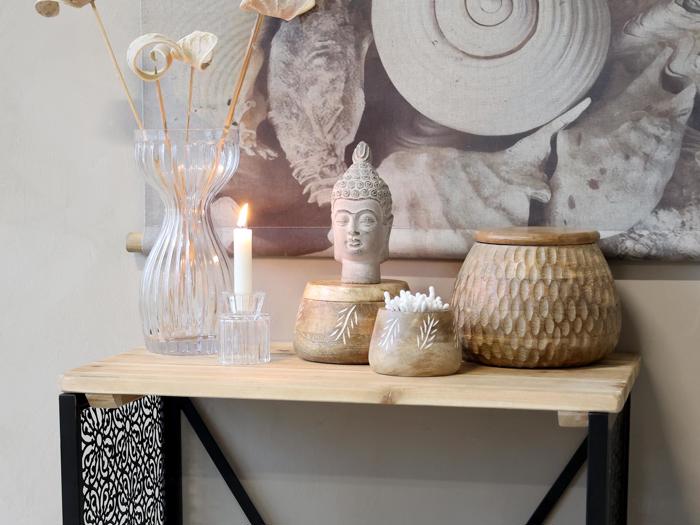 Et træbord med en klar vase med tørrede blomster, et tændt stearinlys, en statue af et buddha-hoved, trækrukker og et tekstureret vægophæng i baggrunden.