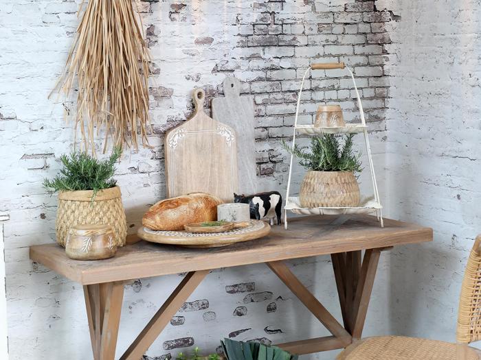 Et rustikt træbord med brød, ost, potteplanter, en sort-hvid ko-figur og dekorative skærebrætter mod en hvid murstensvæg.