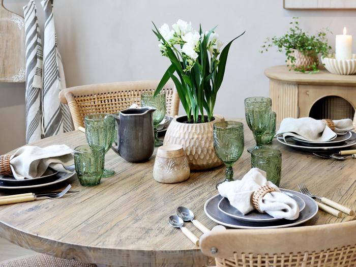 Et rundt træbord dækket til fire med grønne glas, neutrale tallerkener, stofservietter og en vase med hvide blomster som midterstykke. Et stearinlys og en potteplante står i baggrunden.