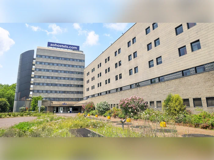 En moderne bygning i flere etager med et aohostels.com-skilt, store vinduer og en anlagt have foran under en blå himmel med skyer.