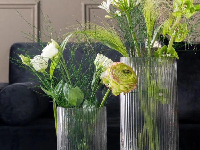 To klare, riflede glasvaser indeholder grønne og hvide blomster med grønne blade på en mørk, sløret baggrund.