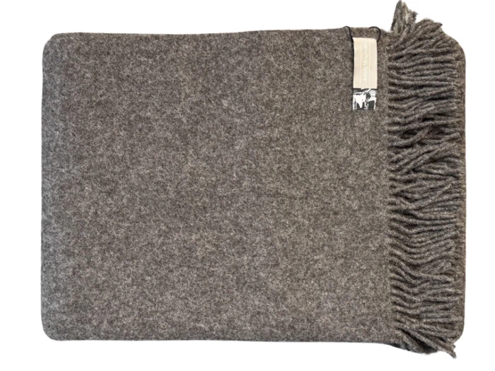 Et gråt uldtæppe med frynser i den ene ende, pænt foldet og med en synlig etiket i øverste højre hjørne.