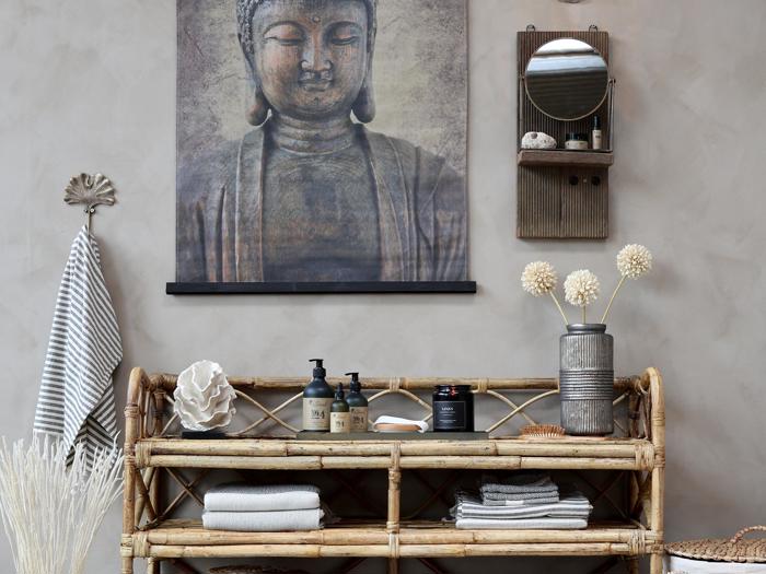 En bambushylde med håndklæder, toiletartikler og dekorationer står under et stort Buddha-kunstværk; et rundt spejl og en væghylde hænger i nærheden og skaber en fredfyldt, spa-agtig atmosfære.