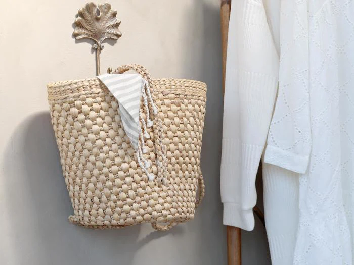 En vævet stråtaske hænger på en dekorativ vægknage formet som et blad, med et hvidt og gråt stribet klæde draperet over tasken. En hvid, struktureret skjorte hænger i nærheden.