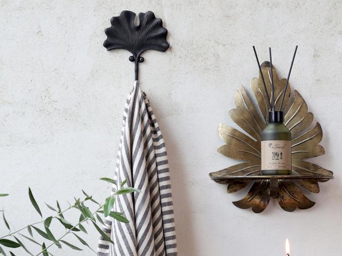 Et stribet håndklæde hænger på en bladformet krog ved siden af en væghylde i bladguld med en diffusorflaske på en lys væg med grønne planter i forgrunden.