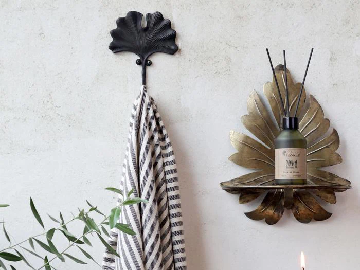 Et stribet håndklæde hænger på en bladformet krog ved siden af en væghylde i bladguld med en diffusorflaske på en lys væg med grønne planter i forgrunden.
