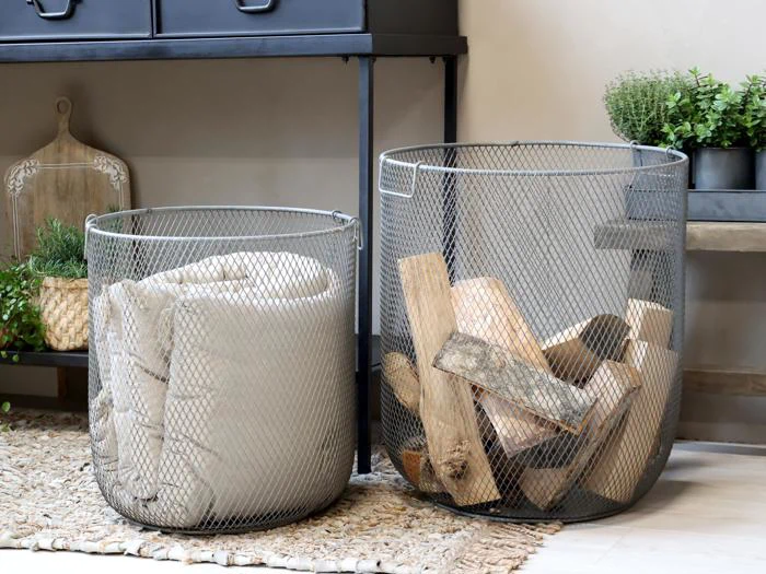 To metalnetkurve står på et vævet tæppe. Den ene kurv indeholder et foldet beige tæppe, mens den anden indeholder brænde. Der er planter og ting til boligindretning i baggrunden.