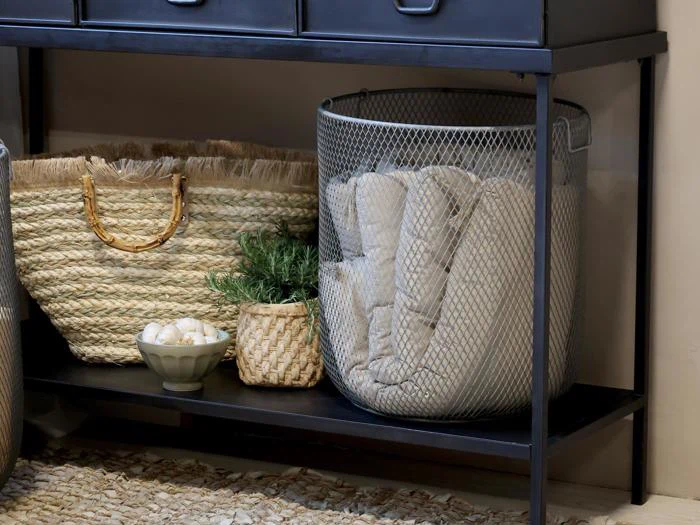 En flettet kurv, en lille potteplante, en skål med skaller og en trådkurv med foldede tæpper er anbragt på en sort metalhylde.