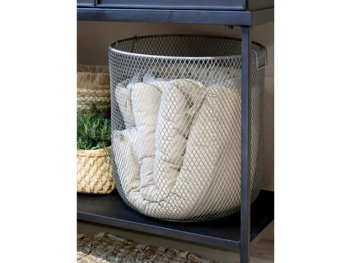 En metalnetkurv med foldede beige tæpper står på en sort hylde ved siden af en flettet kurv med grønne kunstige planter.