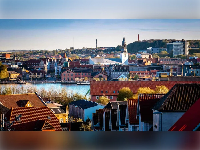 Et levende bybillede med bygninger med røde tage, et fremtrædende kirketårn, træer og vand i baggrunden under en klar blå himmel.