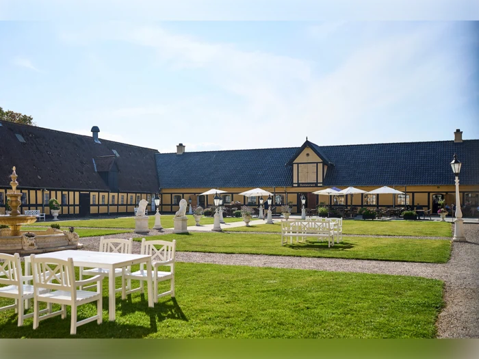 En gårdhave med hvide udendørsborde og -stole, grønt græs, statuer, et springvand og en stor gul bygning i baggrunden under en klar himmel.