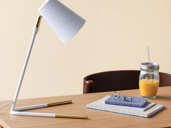 En moderne hvid skrivebordslampe, en stak notesbøger med glas og et syltetøjsglas med appelsinjuice står på et træskrivebord mod en beige baggrund.