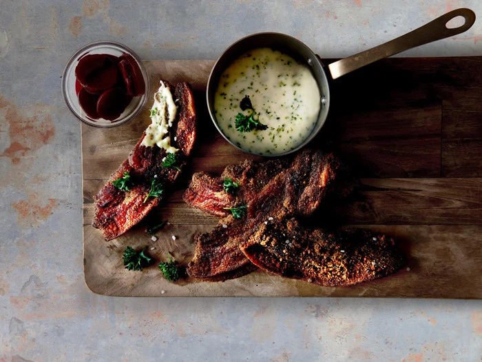 En træplade med krydrede kødstrimler, en lille gryde med cremet sauce, en skål med skiveskårne rødbeder og pyntet med friske krydderurter.