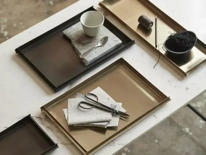 Rektangulære metalbakker på et bord med en kop, en ske, en saks, servietter, en trådspole og en skål med mørkt garn.