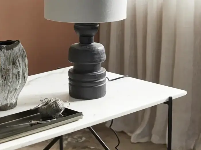 Et moderne hvidt bord med en mørk skulpturel lampe, en keramikvase og en bakke med garn og saks. Lyse gardiner hænger i baggrunden.