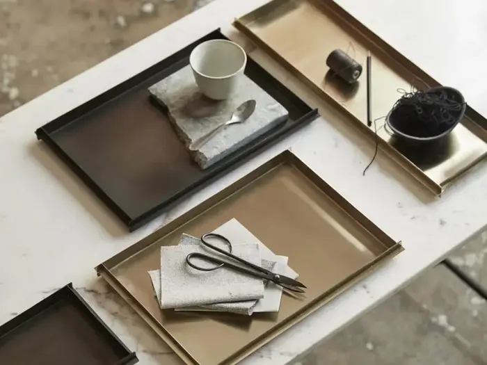 Et hvidt bord med metalbakker med forskellige ting: en kop og en ske på en serviet, tråd og en skål og en saks på sammenfoldede klude.