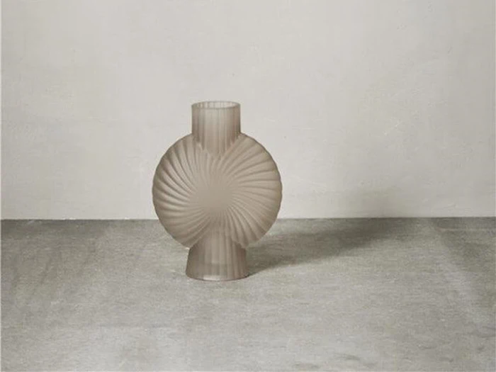 En rund, riflet, gennemskinnelig beige vase med smal hals og bund står på et gråt betongulv mod en almindelig hvid væg.