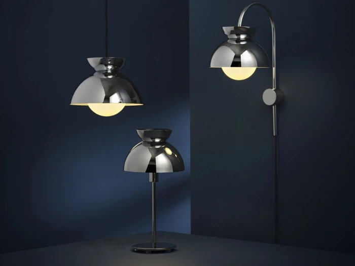 Tre moderne kromlamper – en pendel, en bordlampe og en væglampe – udstråler varmt lys på en mørkeblå baggrund og fremhæver de slanke, reflekterende metaldesign.