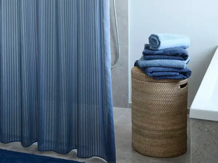 En flettet vasketøjskurv med sirligt foldede blå håndklæder står ved siden af et badekar og et blåt bruseforhæng på et badeværelse.