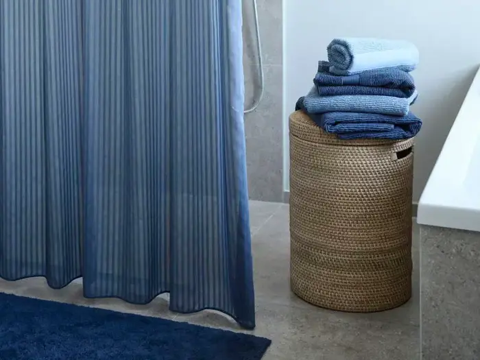 En flettet vasketøjskurv med sirligt foldede blå håndklæder står ved siden af et badekar med et blåt badeforhæng og en matchende bademåtte i nærheden.