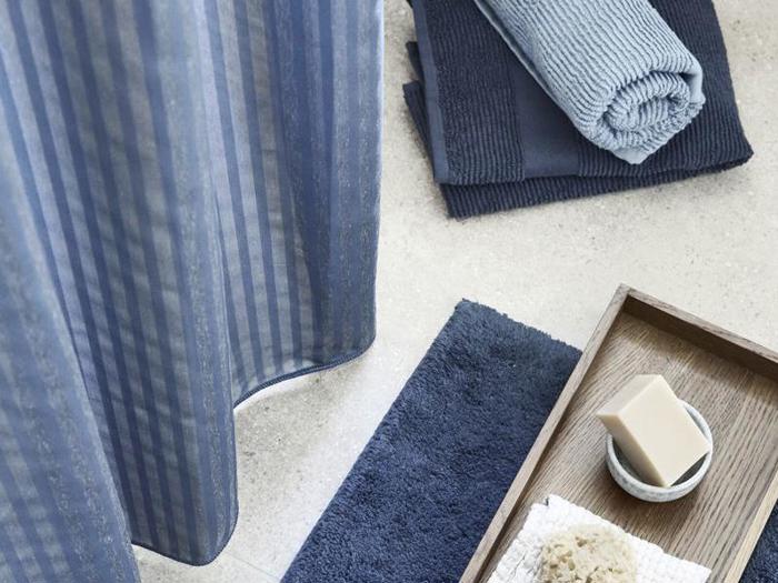 Blå og marineblå håndklæder, et blåstribet badeforhæng, en blå bademåtte og en træbakke med et stykke sæbe og en svamp på et lyst badeværelsesgulv.