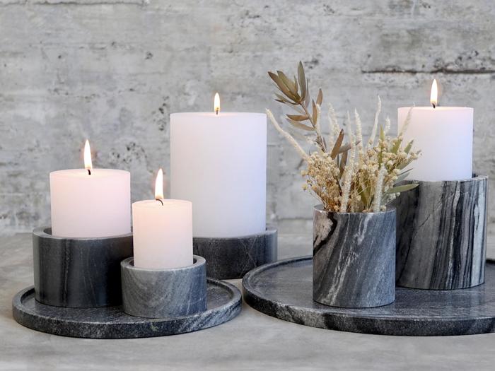 Fire hvide lys i forskellige højder brænder i runde, mørke marmorstager ved siden af en matchende vase med tørret græs, alt sammen placeret på stenbakker mod en grå struktureret baggrund.
