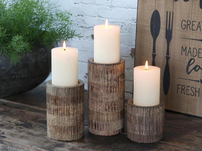 Tre hvide stearinlys er tændt og placeret på rustikke træholdere i forskellige højder på et træbord ved siden af en grøn plante og et dekorativt køkkenskilt.