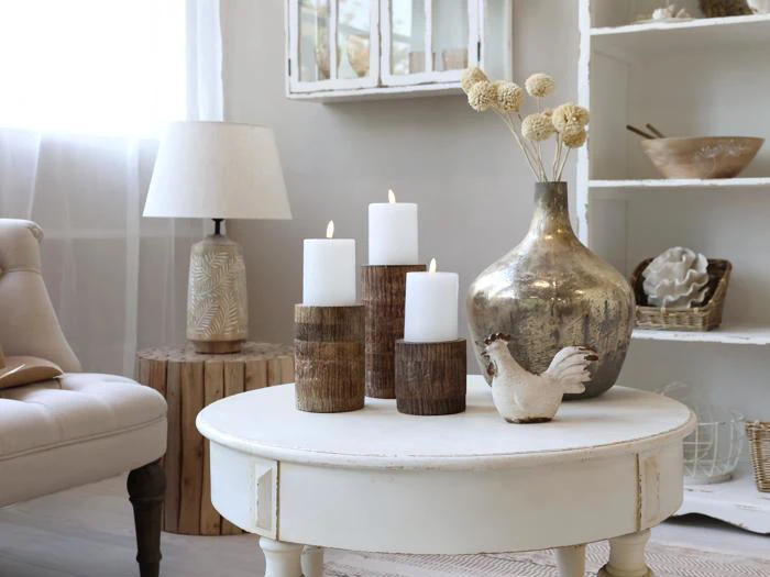 En hyggelig stue med et hvidt rundt bord med tre tændte bloklys, en metalvase med tørrede blomster og en keramisk kylling. Neutrale toner og naturligt lys fylder rummet.