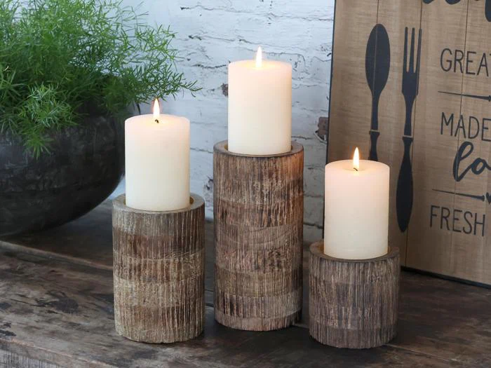 Tre hvide stearinlys er tændt og placeret på rustikke træholdere i forskellige højder på et træbord ved siden af en grøn plante og et dekorativt køkkenskilt.